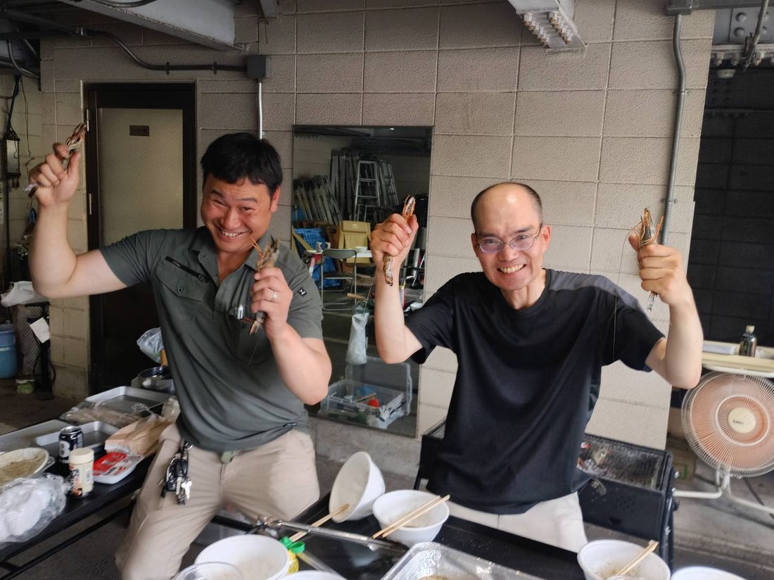 社内バーベキューの様子。事務所前のガレージで、社員2名が串に刺した大きな海老を掲げて笑っている