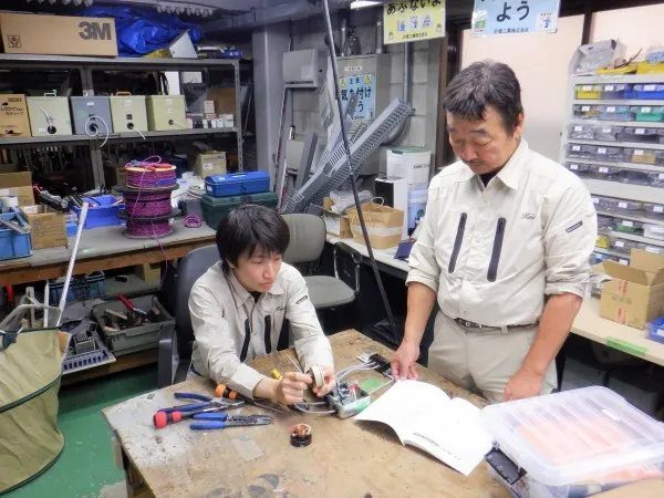 作業場の作業台で、若手社員が配線作業をし、先輩社員が図面を見ながら教えている様子