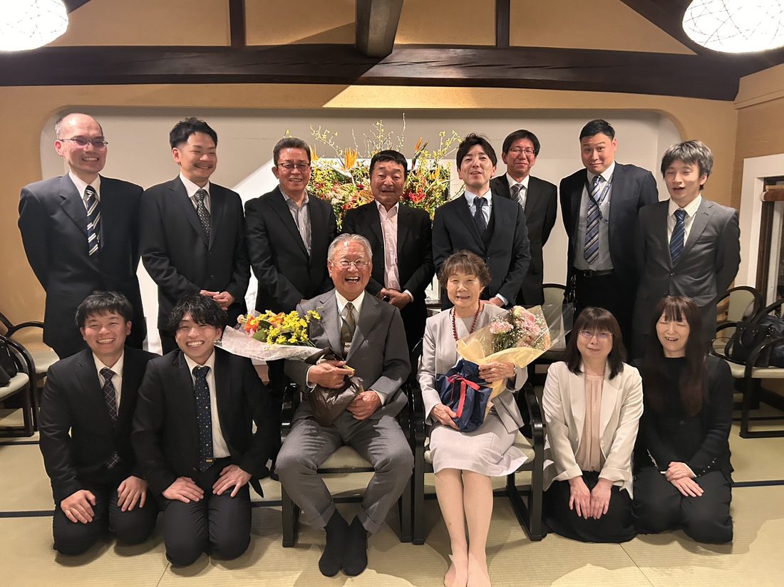 前社長の退任慰労会での集合写真。花束を持った前社長夫妻を囲んで社員が並んでいる