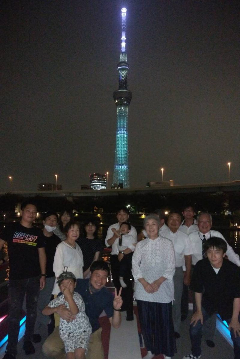 貸切の屋形船の甲板での集合写真。ライトアップされた東京スカイツリーを背に社員と家族が並んでいる